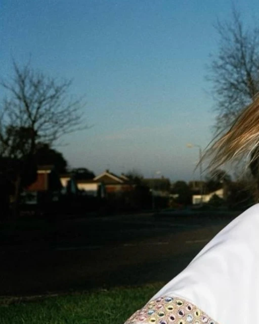 Blonde hair woman's shoulder with ornamental sleeve, road and houses in soft focus behind, blue sky. 