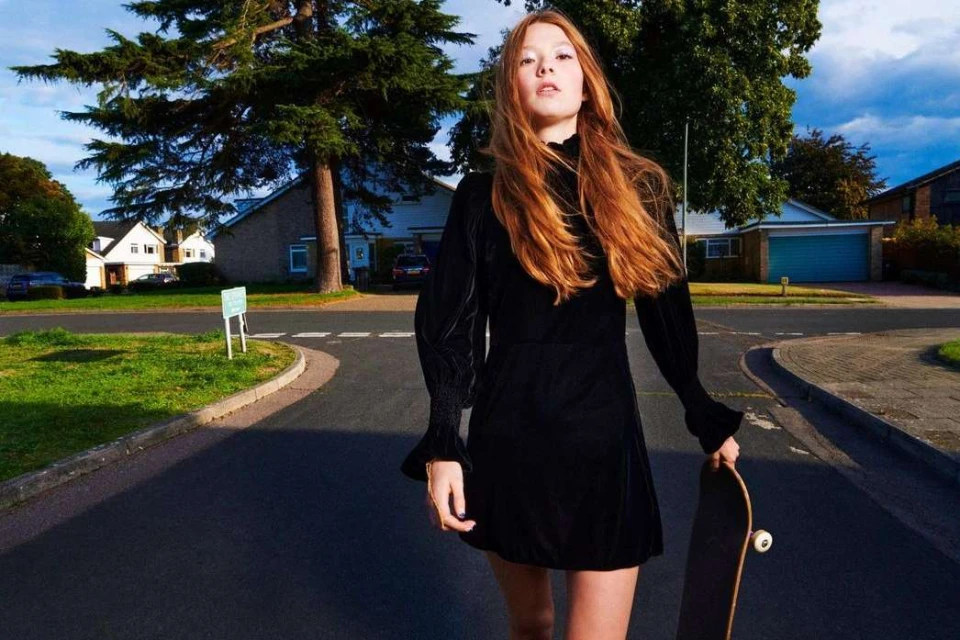 Young woman in a black dress walks on a suburban street, holding a skateboard, with houses and trees in the background on a sunny day.