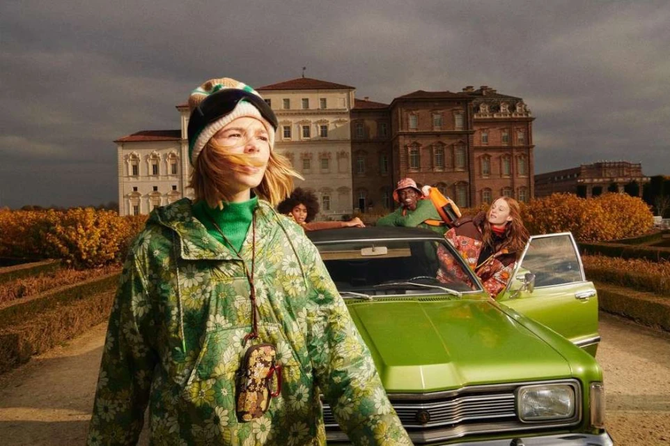 Fashionable woman in floral jacket stands near a green vintage car with group in background, set against building and cloudy sky.