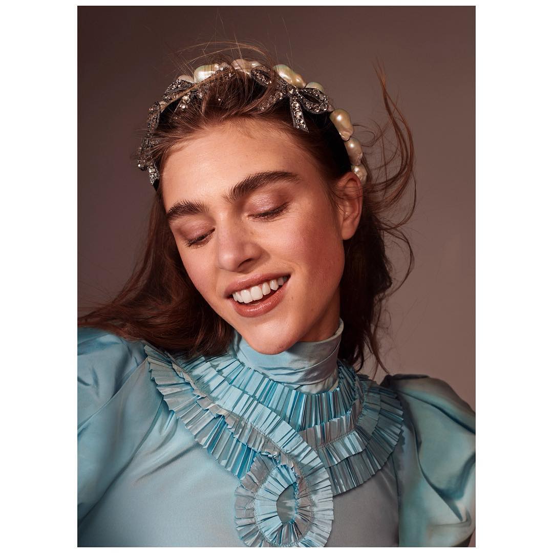 Close-up of a smiling adult woman wearing a pearl headband, a light blue blouse with ruffles, and light makeup indoors.