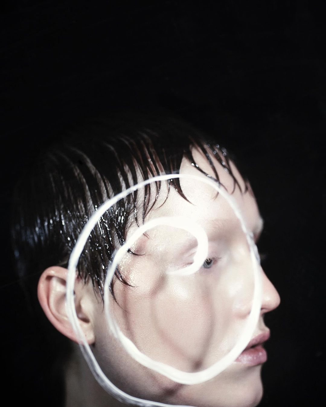 Headshot of a person with short, wet hair and a white, spiraled, painted design over their face, set against a stark black background.