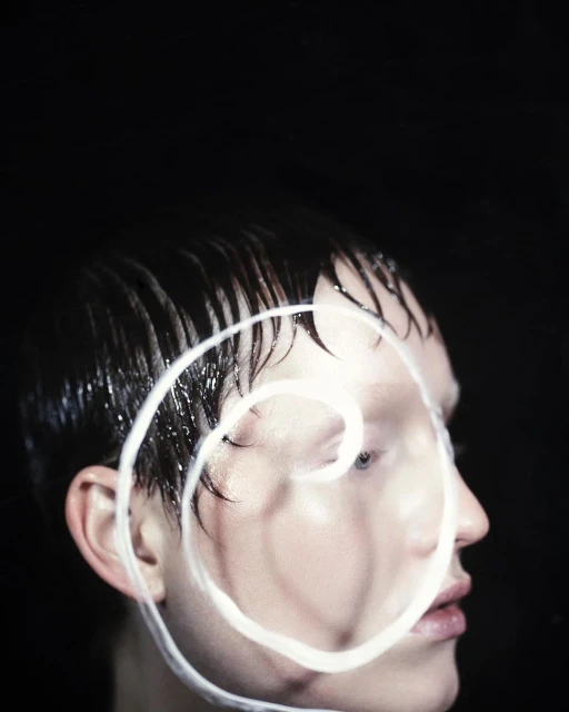 Headshot of a person with short, wet hair and a white, spiraled, painted design over their face, set against a stark black background.