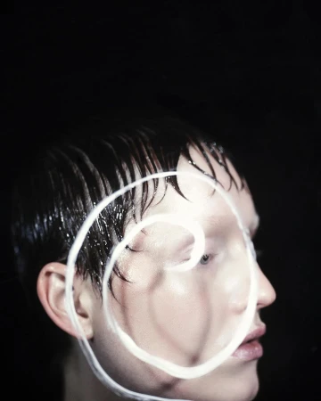 Headshot of a person with short, wet hair and a white, spiraled, painted design over their face, set against a stark black background.
