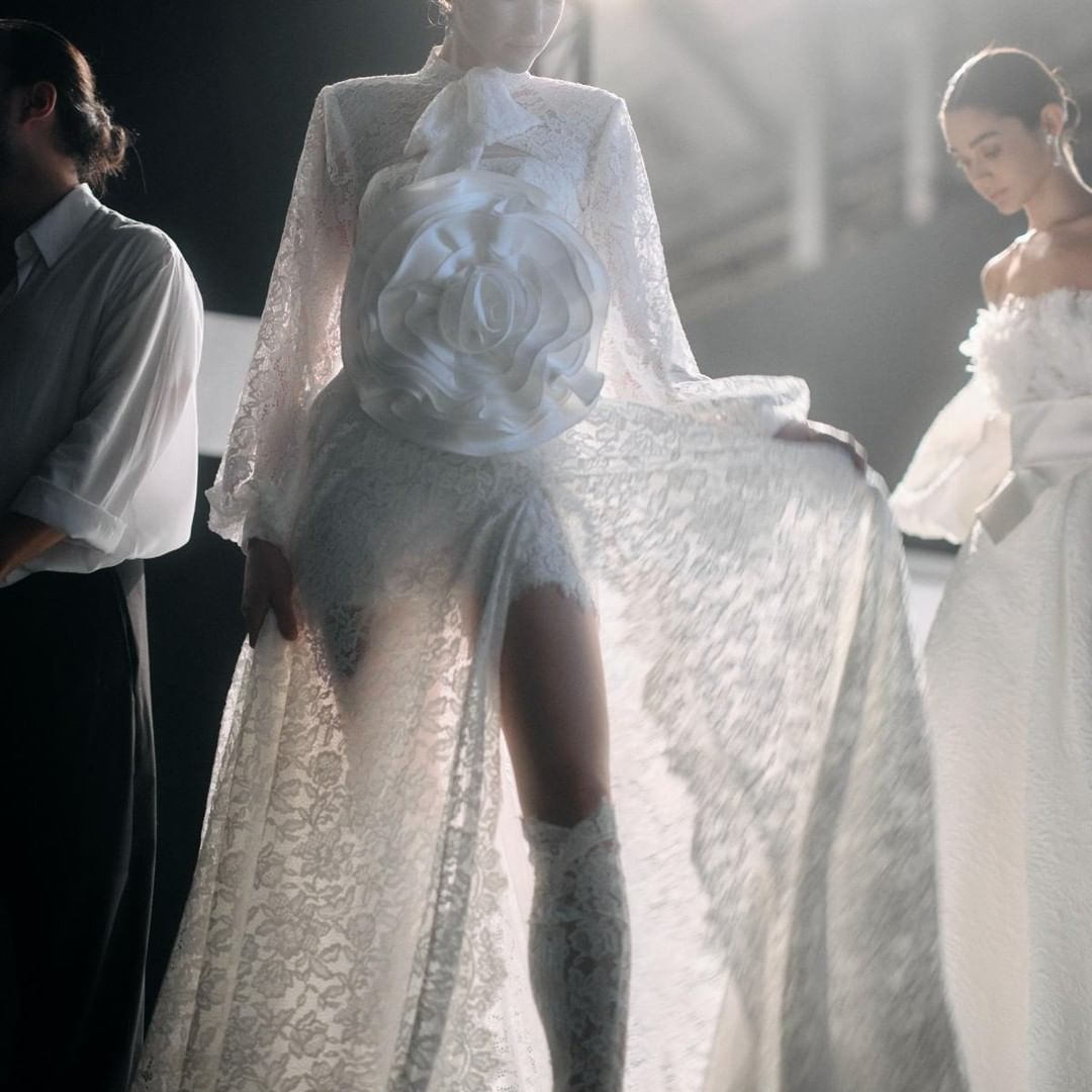 A model in a white lace gown with a large floral accent poses alongside two other figures in a fashion setting with soft lighting.