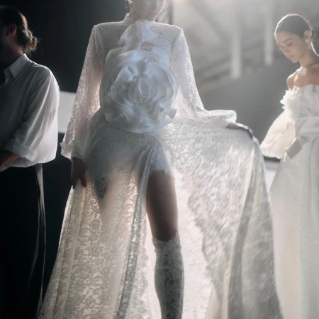 A model in a white lace gown with a large floral accent poses alongside two other figures in a fashion setting with soft lighting.