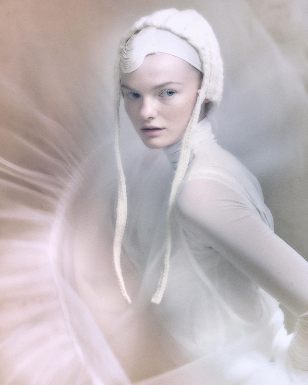 Close-up portrait of a fair-skinned adult woman with blue eyes, wearing a white knit hood and sheer dress against a blurred background.