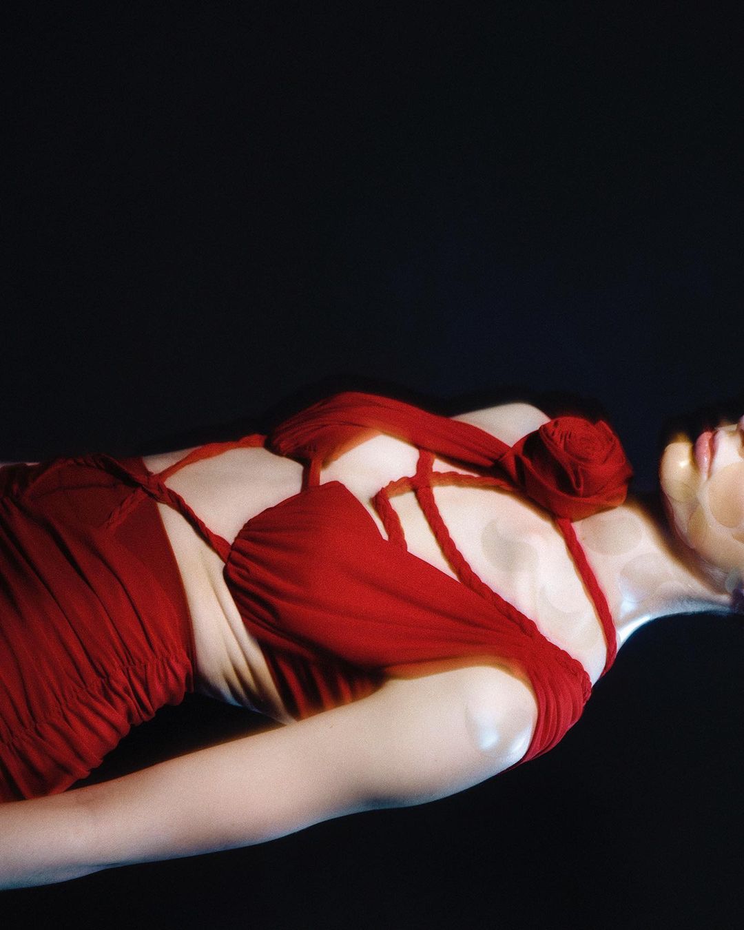 Adult female lying down wearing red draped dress against black background in studio lighting. Beauty and fashion editorial photograph.