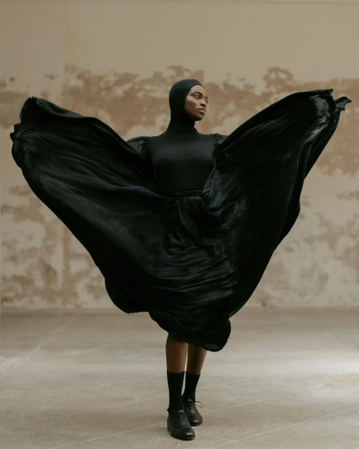 Elegant Black woman posing in a draped black dress, dancing in a light colored room in a fashion shot showcasing movement and style.
