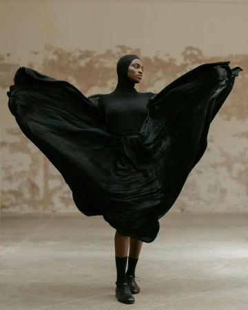 Elegant Black woman posing in a draped black dress, dancing in a light colored room in a fashion shot showcasing movement and style.
