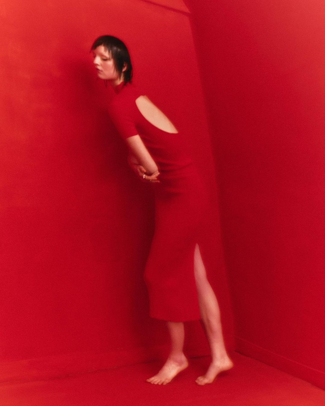 Woman in red dress poses against a red wall, showcasing the dress's backless design and the minimalist color palette.