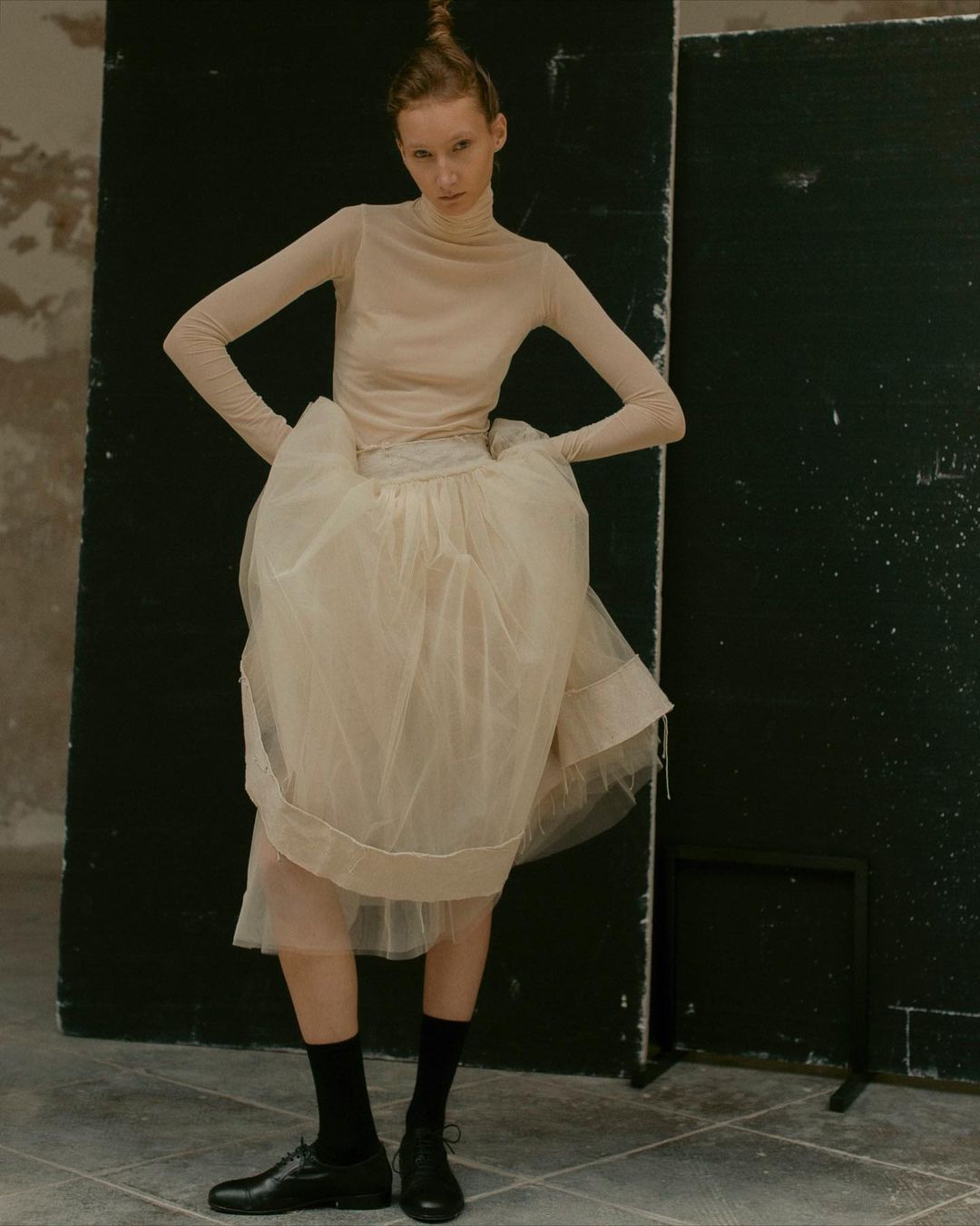 A slender fashion model strikes a pose indoors, wearing a beige long-sleeve top and a large, layered tulle skirt with black socks and leather shoes.