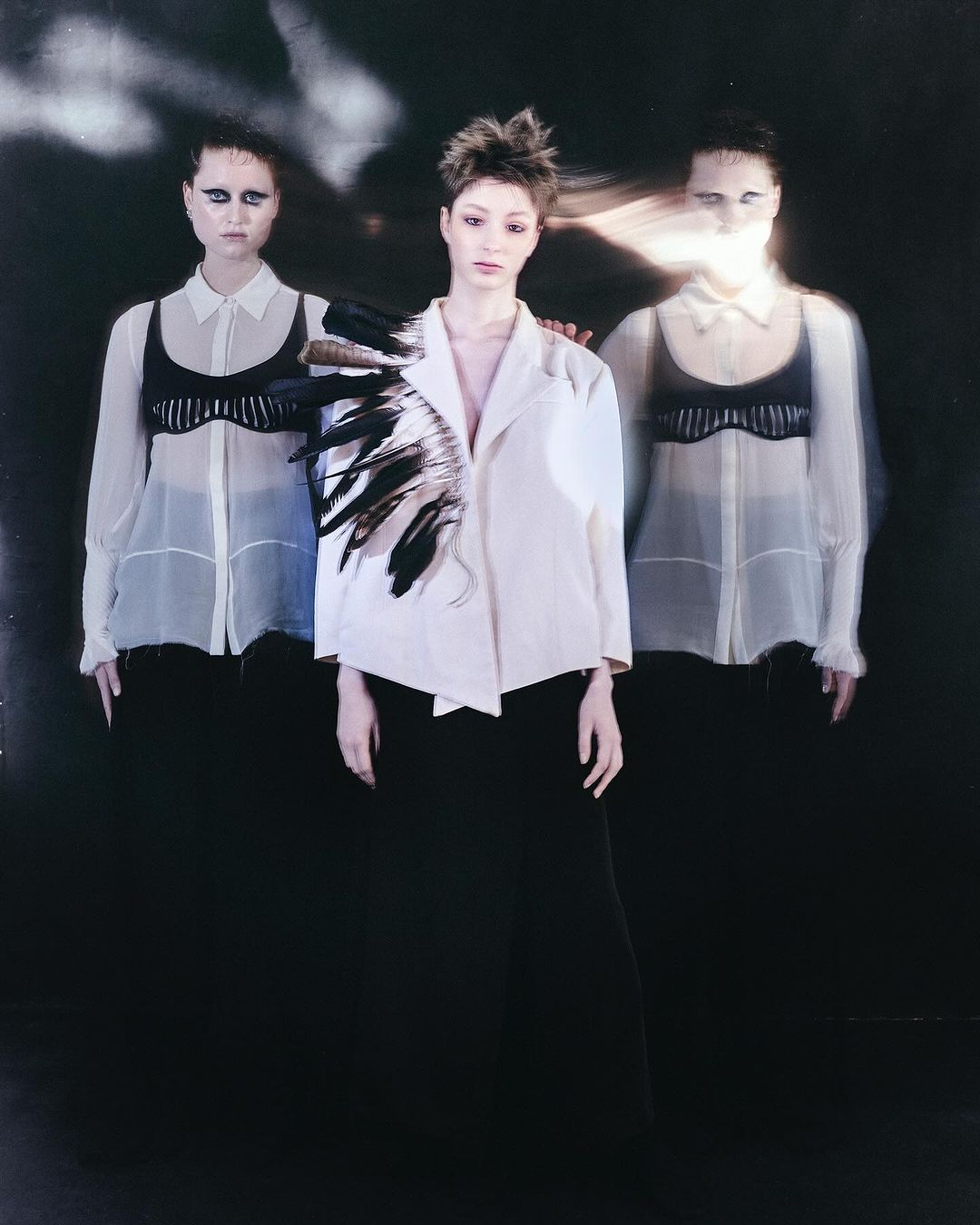 Three women pose in a dimly lit studio. The central figure wears a white jacket with black feathers on one shoulder, creating an artistic and striking image.