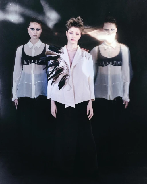 Three women pose in a dimly lit studio. The central figure wears a white jacket with black feathers on one shoulder, creating an artistic and striking image.