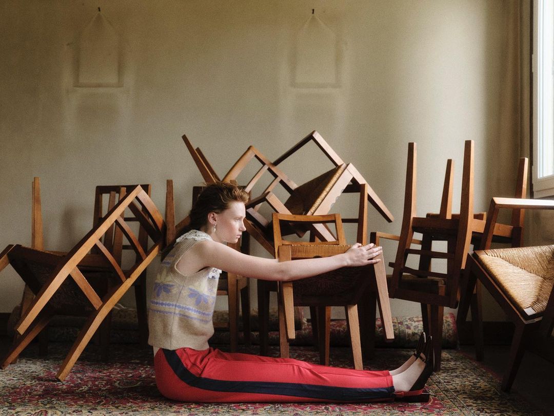 Woman in retro tracksuit sits amongst stack of wooden chairs inside a vintage room, with soft natural light illuminating the scene.