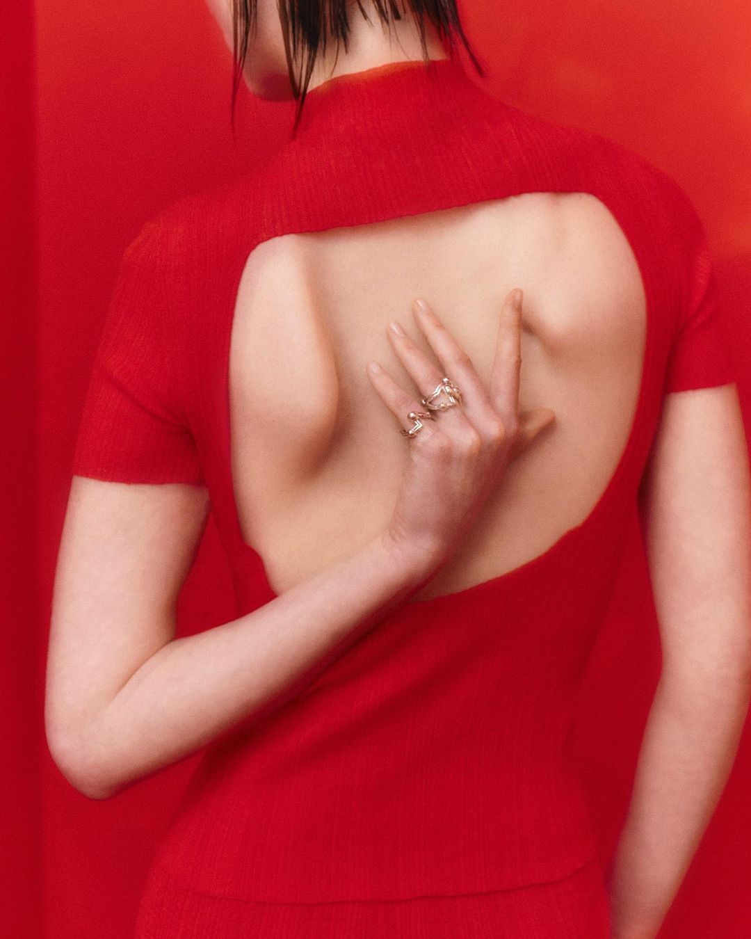 A slender woman wearing a red outfit and striking jewelry poses against a solid red background in a captivating fashion photograph.
