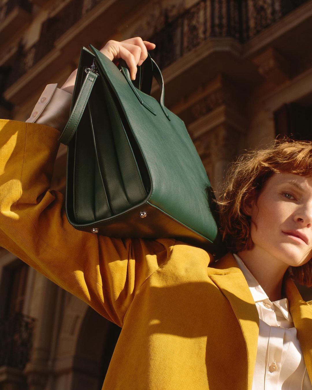 Woman in yellow jacket holds a dark green tote bag in front of a building exterior, bathed in warm sunlight.