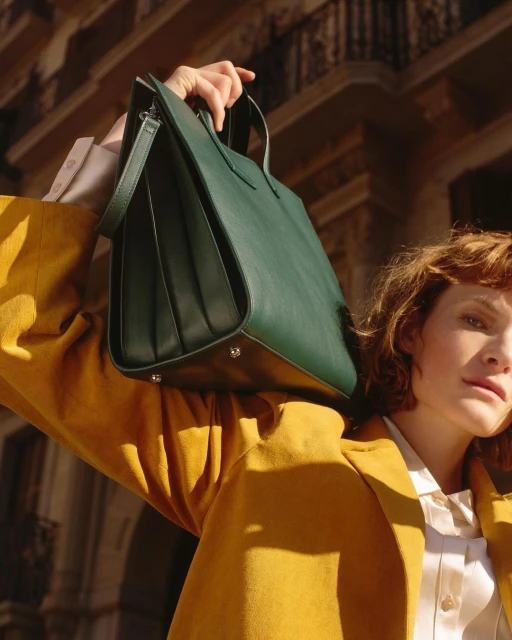Woman in yellow jacket holds a dark green tote bag in front of a building exterior, bathed in warm sunlight.