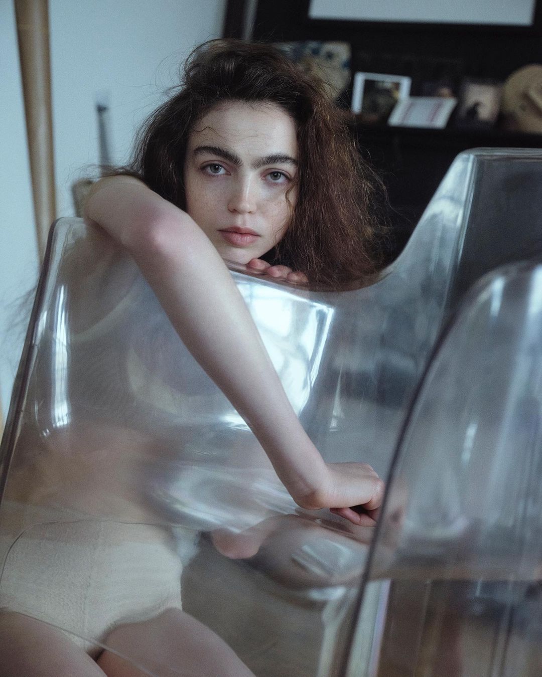 A young adult woman with brown wavy hair gazes at the camera while seated in a transparent chair indoors with soft, diffused lighting.