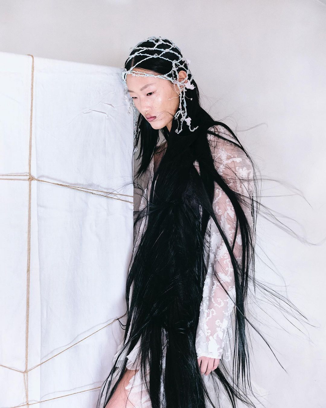 Asian woman with long black hair wearing a white lace dress and beaded headpiece stands next to a wrapped white cube in studio light.