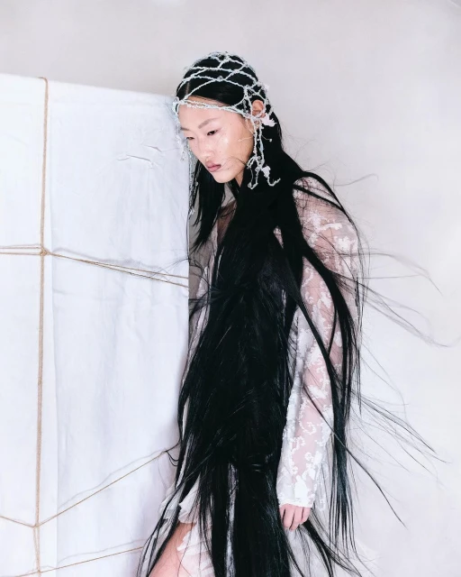 Asian woman with long black hair wearing a white lace dress and beaded headpiece stands next to a wrapped white cube in studio light.