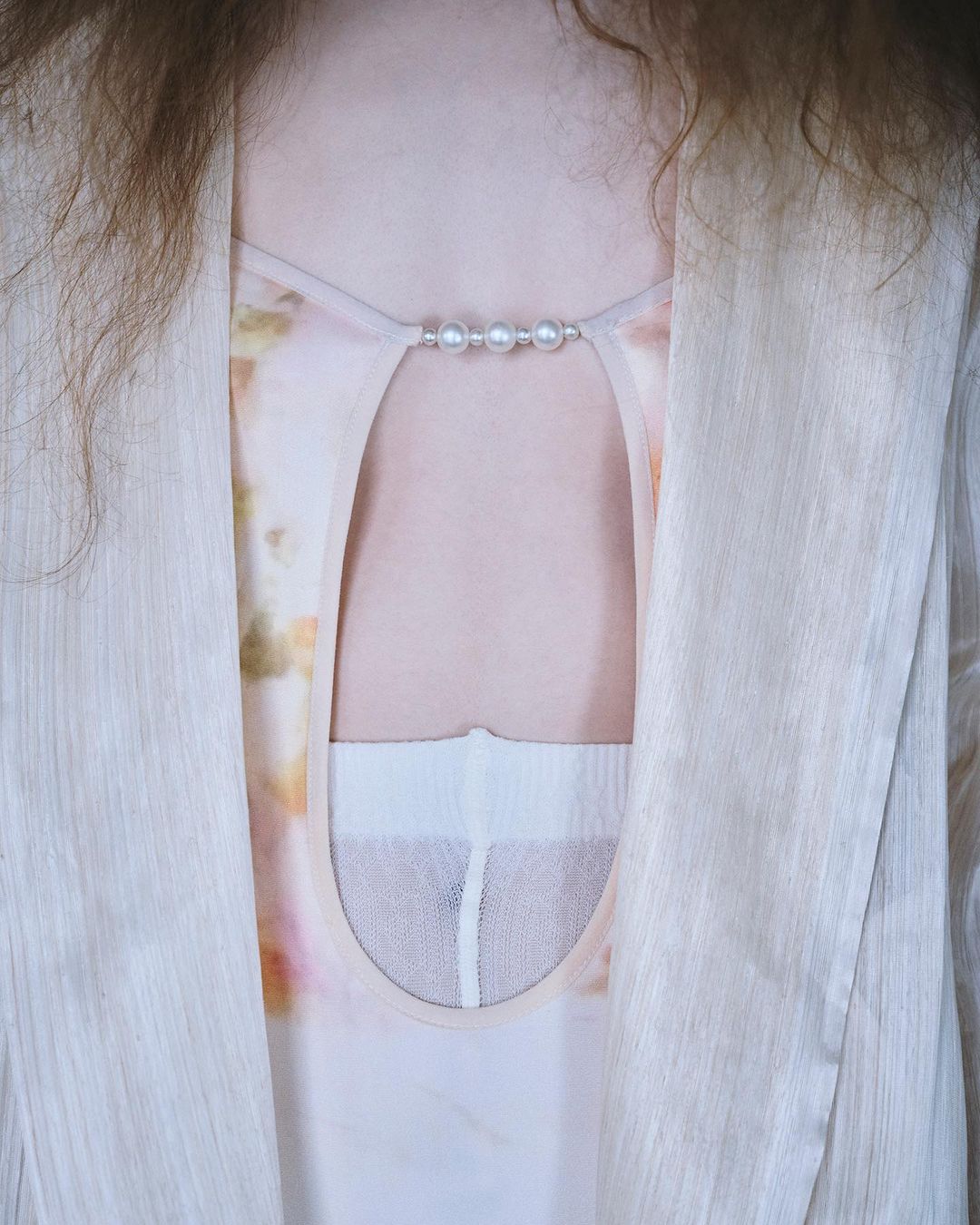 Woman's torso with pearl-detailed top, sheer cardigan, and open back in a brightly lit, neutral indoor setting. Fashion forward styling.