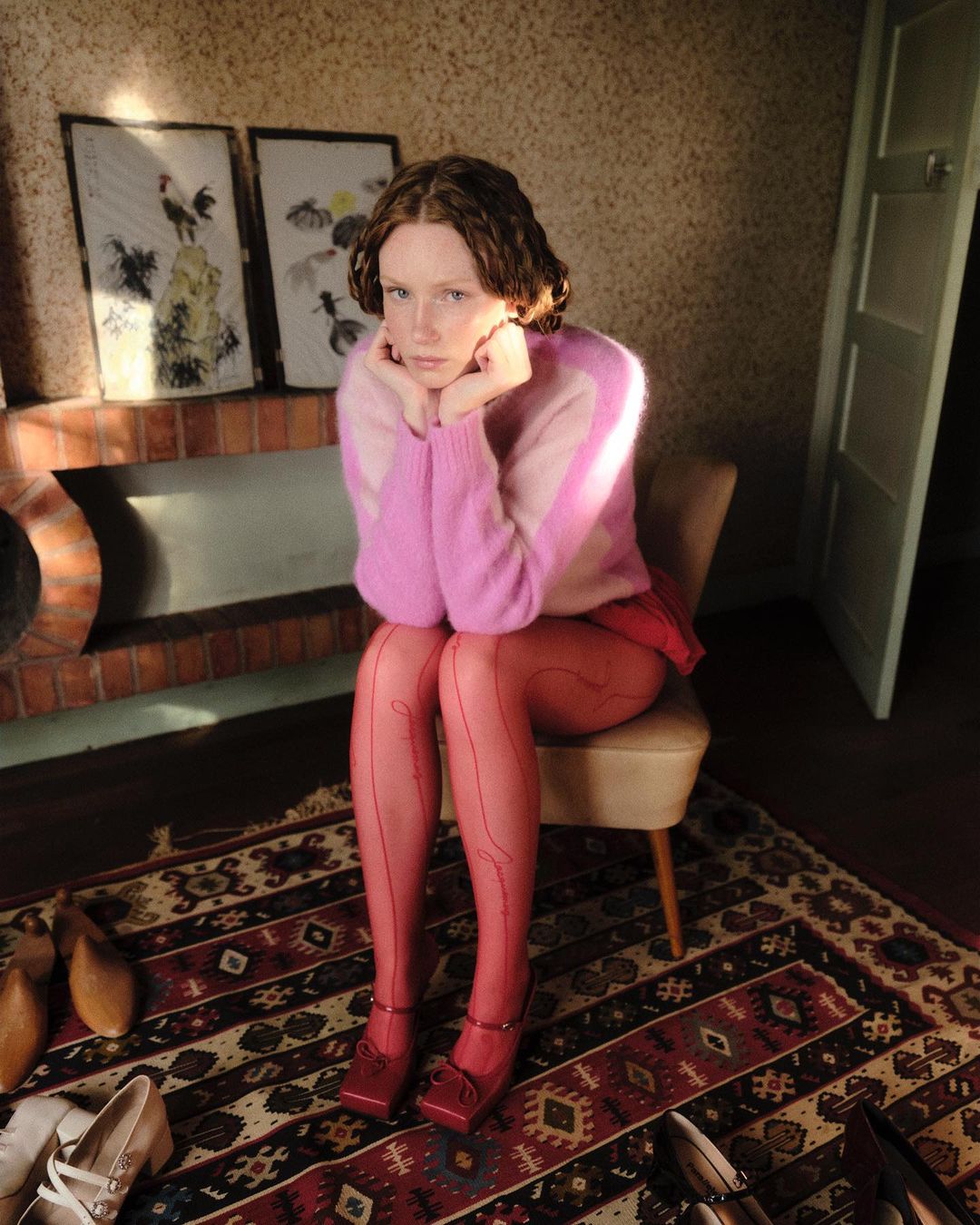 A redhead woman sits indoors on a chair, wearing a pink sweater, tights, and heels, with shoes scattered around her on a patterned rug.