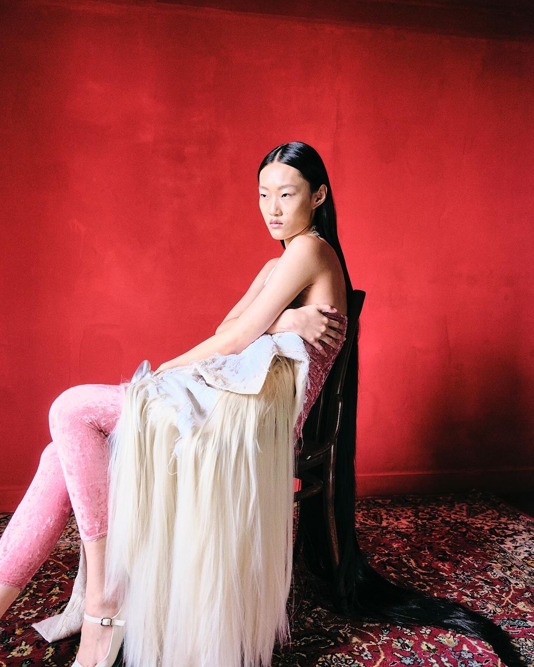 Asian woman seated in a chair with an unusual garment on a patterned rug in front of a red textured wall, fashion shot.