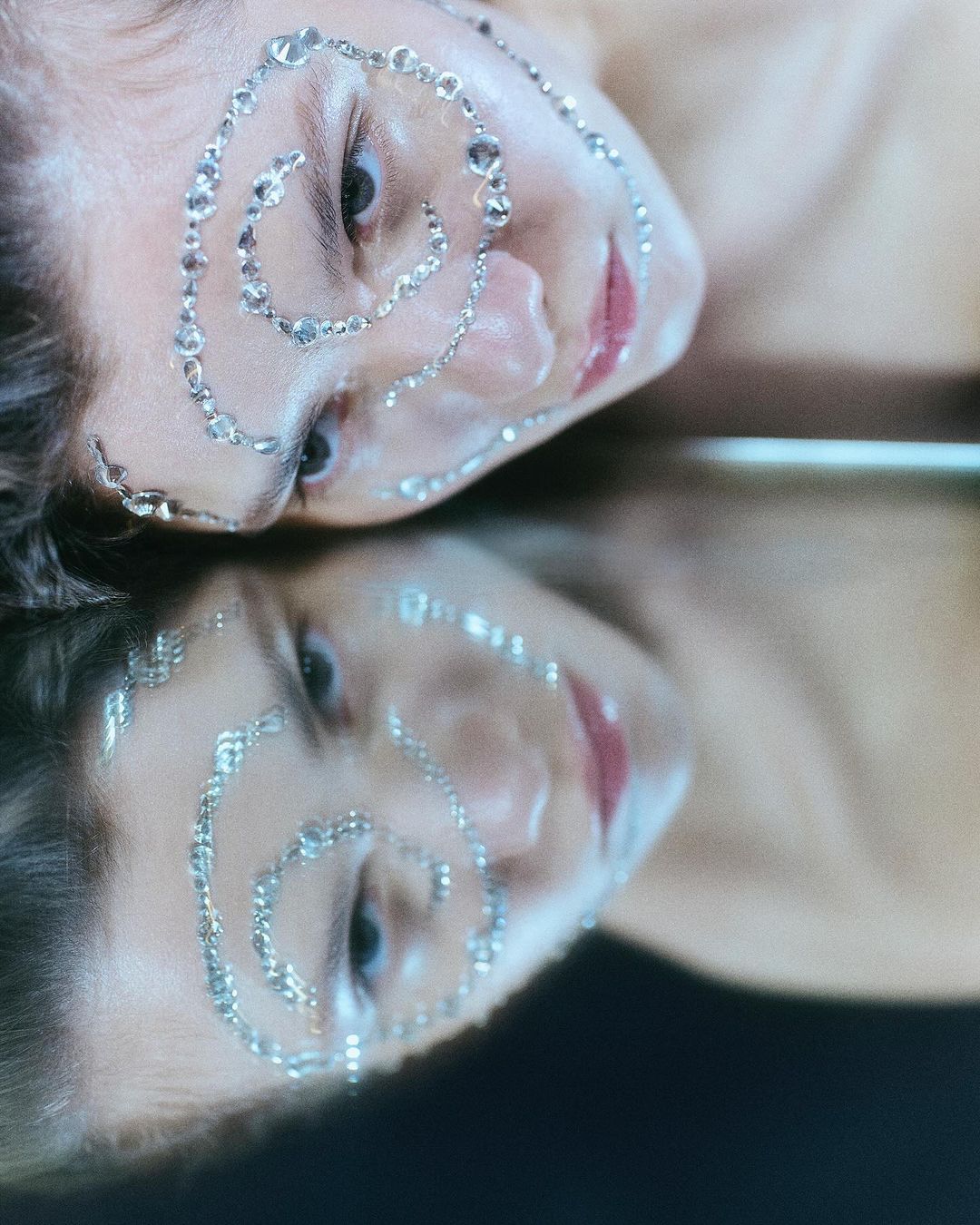 Close-up studio shot of a young woman with silver rhinestones spiraling around her face, reflected in a mirror surface.