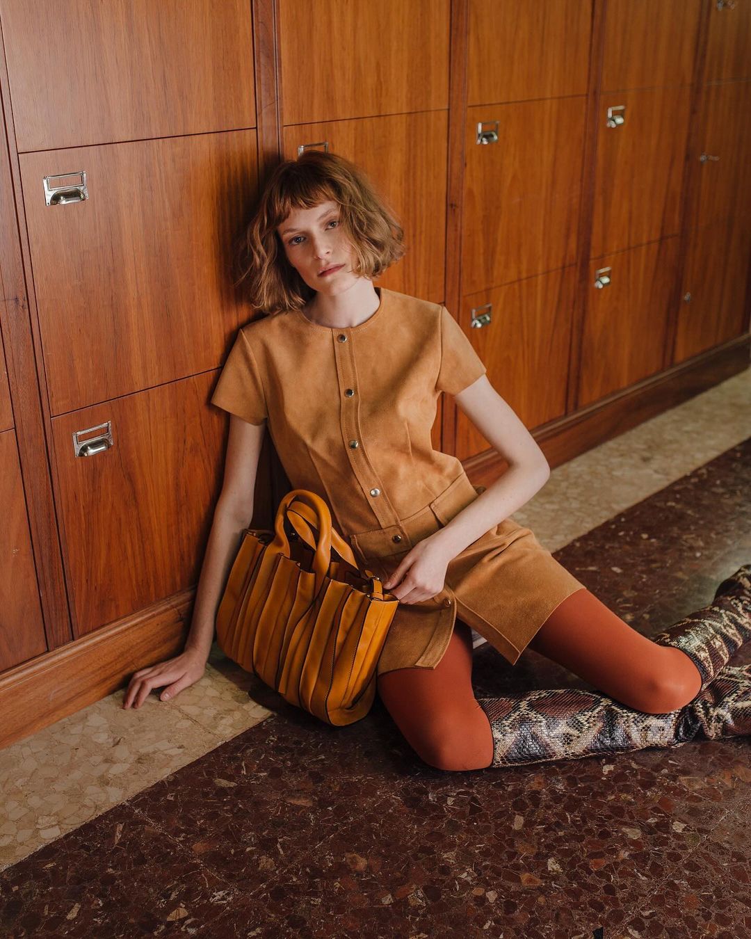Indoor fashion shot of a woman in an ochre short dress and patterned boots sitting on a marble floor with a leather tote bag.