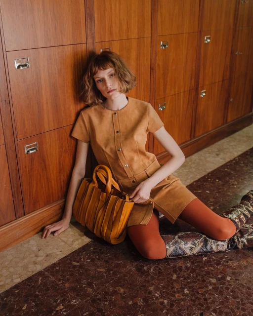 Indoor fashion shot of a woman in an ochre short dress and patterned boots sitting on a marble floor with a leather tote bag.