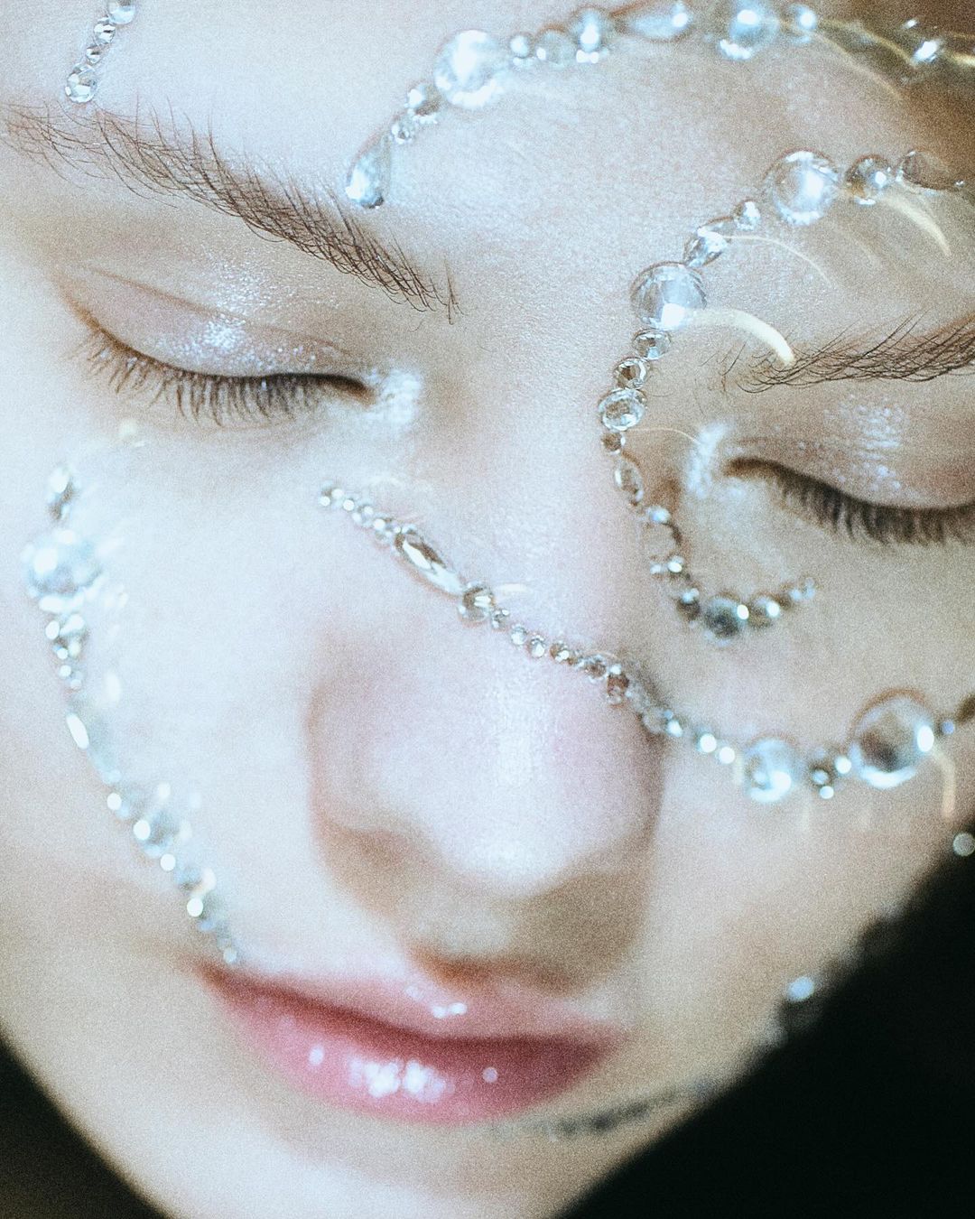 Close-up portrait of a woman with closed eyes and glass-bead embellishments, showcasing a beauty or skincare aesthetic. Soft, diffused lighting.