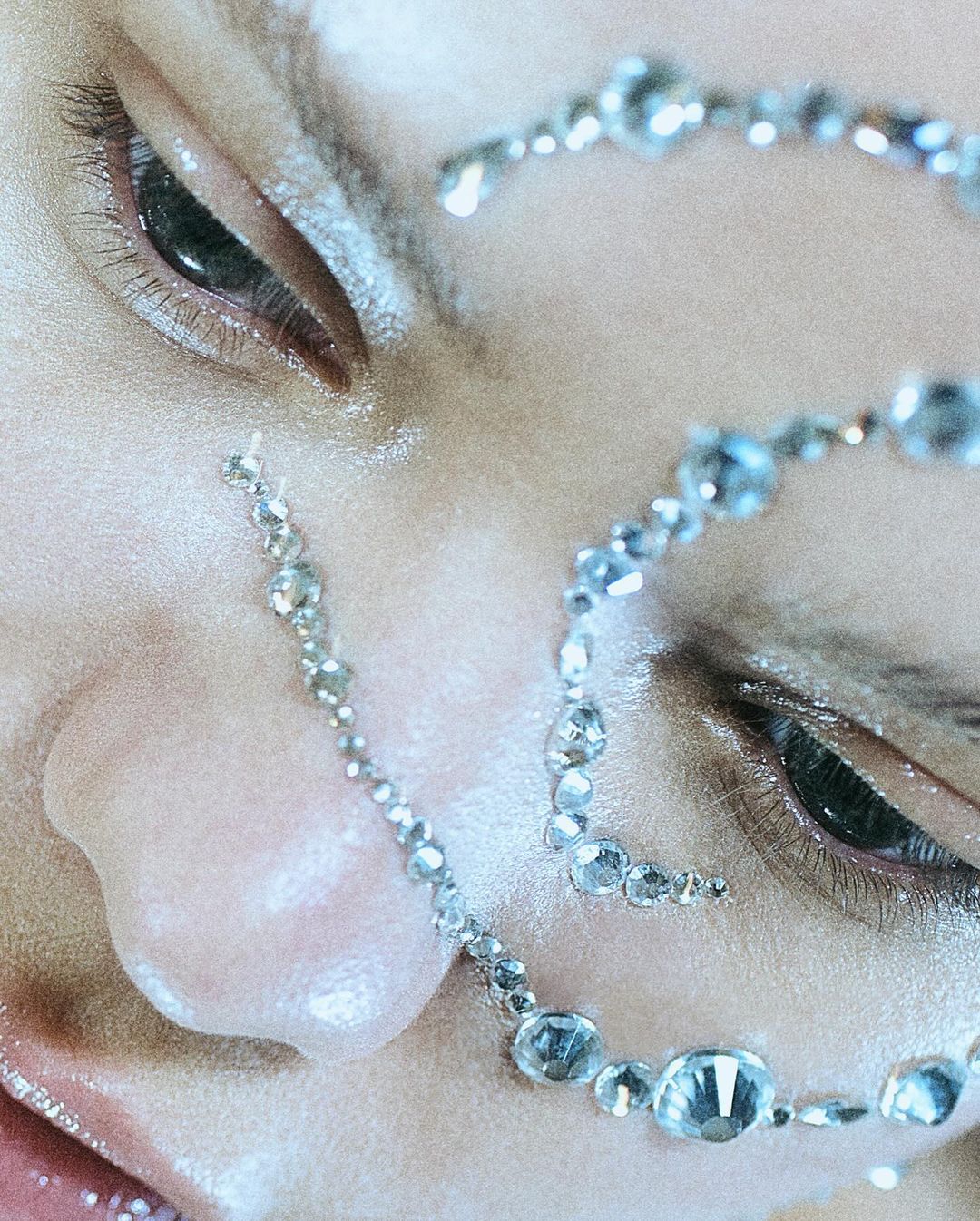 Close-up beauty shot of a woman's face adorned with strands of gem-like beads, emphasizing her eyes and facial features in a light, airy setting.