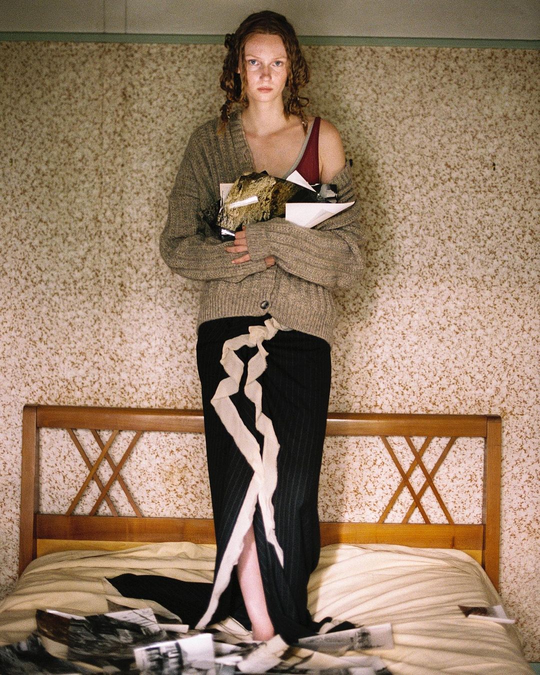 A woman stands on a bed in front of a wallpapered wall holding books, wearing a cardigan and a skirt with ribbons, surrounded by photos.