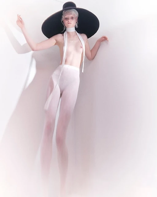 A woman with white hair poses in front of a white wall, wearing sheer pants, a neck scarf, and a large black sun hat.