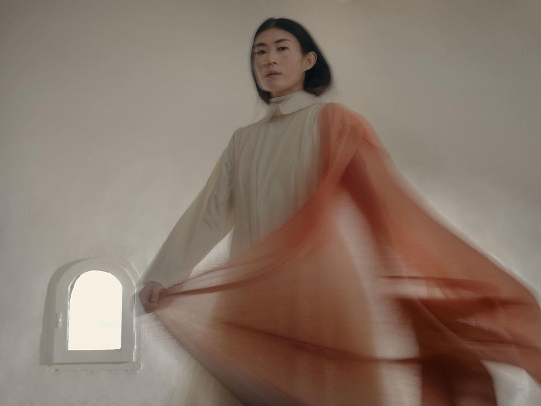 Woman in white dress poses indoors with a flowing orange fabric. Light, soft focus, and neutral tones create a serene fashion editorial.
