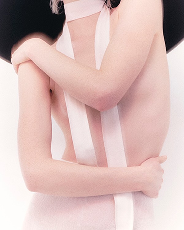 Close-up portrait of a woman wearing a pale pink choker with long trailing ribbons and a wide-brimmed black hat against a white background