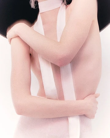 Close-up portrait of a woman wearing a pale pink choker with long trailing ribbons and a wide-brimmed black hat against a white background