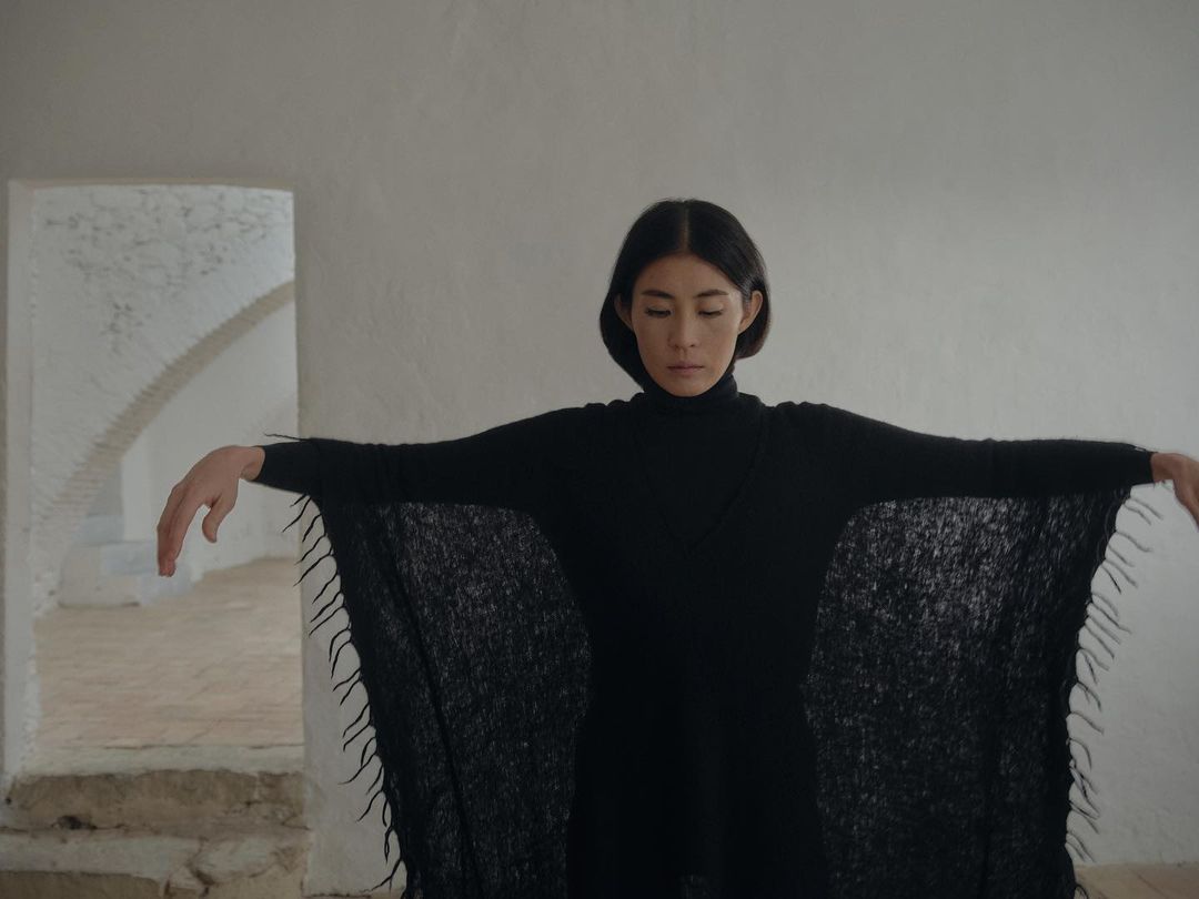 Woman in black turtleneck and wide-sleeved sweater stands in bright white room with arched doorway and textured walls.