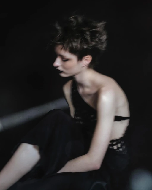 Portrait of a person with spiky hair and dark lipstick, wearing a black dress in a dark studio with soft lighting.