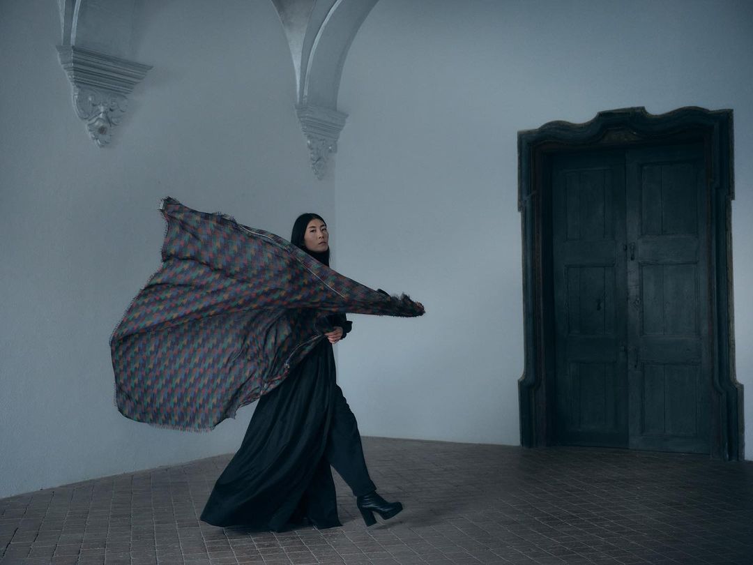 A woman in a flowing coat walks in a stark, old room with dark wood door and white walls, creating a dramatic fashion editorial image.