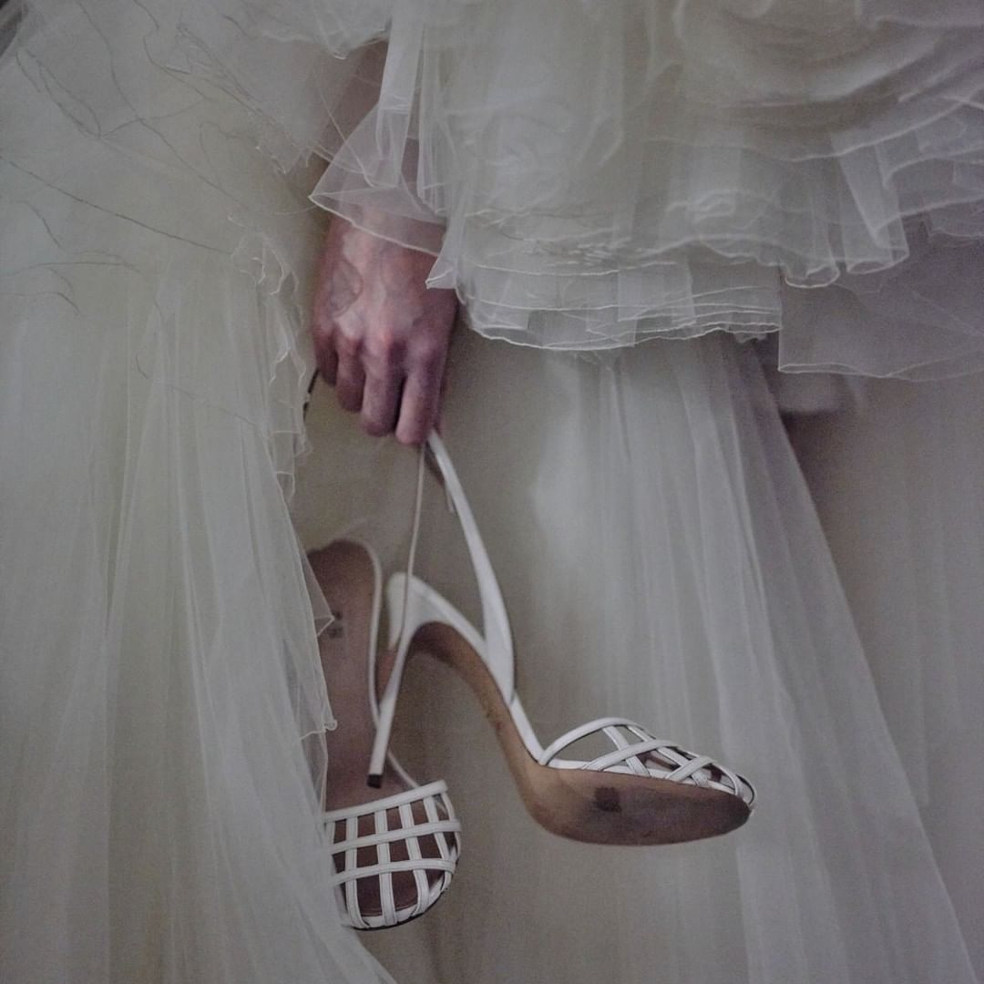 Close-up on an adult's hand holding a pair of white high-heeled shoes, a white wedding dress is in soft focus in the background.