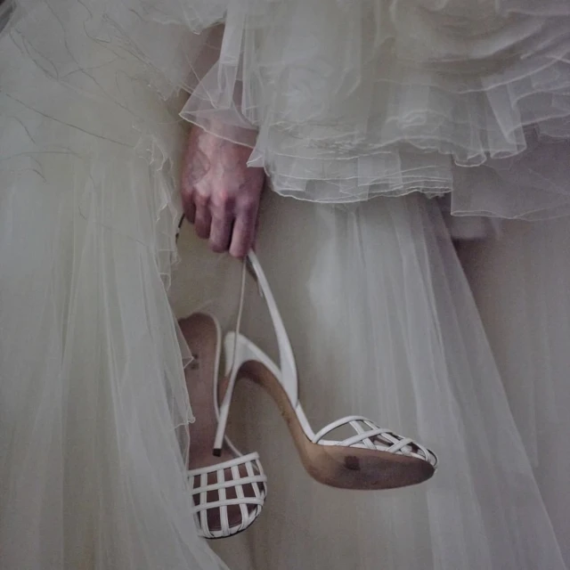 Close-up on an adult's hand holding a pair of white high-heeled shoes, a white wedding dress is in soft focus in the background.
