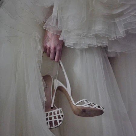 Close-up on an adult's hand holding a pair of white high-heeled shoes, a white wedding dress is in soft focus in the background.