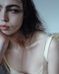 Close-up portrait of a young woman with dark hair and freckles resting her head on her hand, wearing a pale sleeveless top.