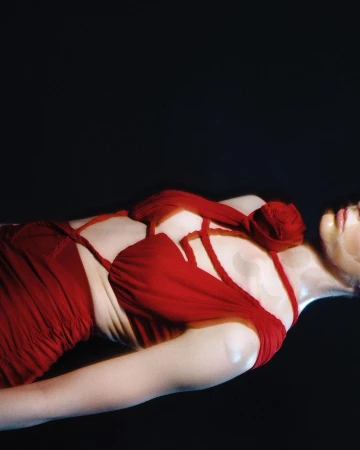 Close-up profile of a woman wearing a red strappy dress with a large fabric rose detail against a dark background, by Eleonore Tornev.