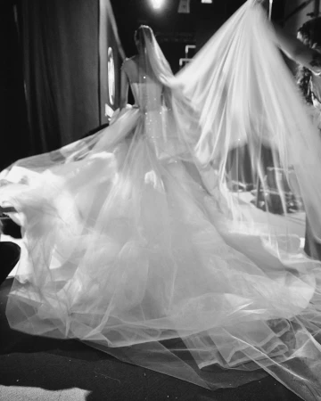 Model seen from behind wearing an elaborate bridal gown with a massive tulle skirt and long veil, photographed in black and white.