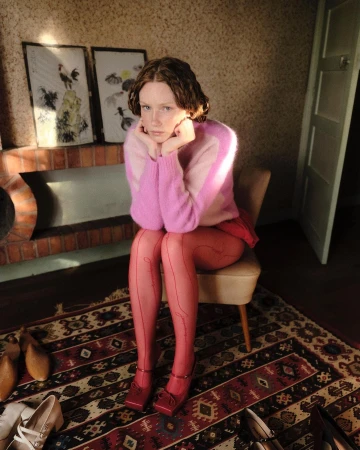 Young woman in a pink mohair sweater and red tights seated on a chair in a domestic interior by Eléonore Tornev