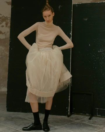 Model in sheer beige turtleneck and voluminous tulle skirt with black socks and shoes posed against dark panels
