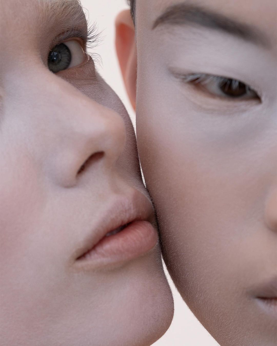 Close-up portrait of two young adult women with pale foundation, pressed together in a beauty or skincare campaign with soft studio lighting.