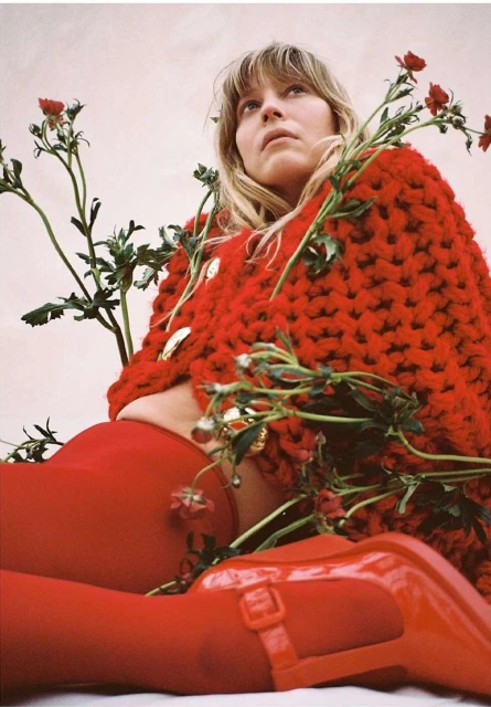 Woman in red knitwear and matching shoes sitting among roses, posing for fashion portrait, shot at eye-level, warm color palette.